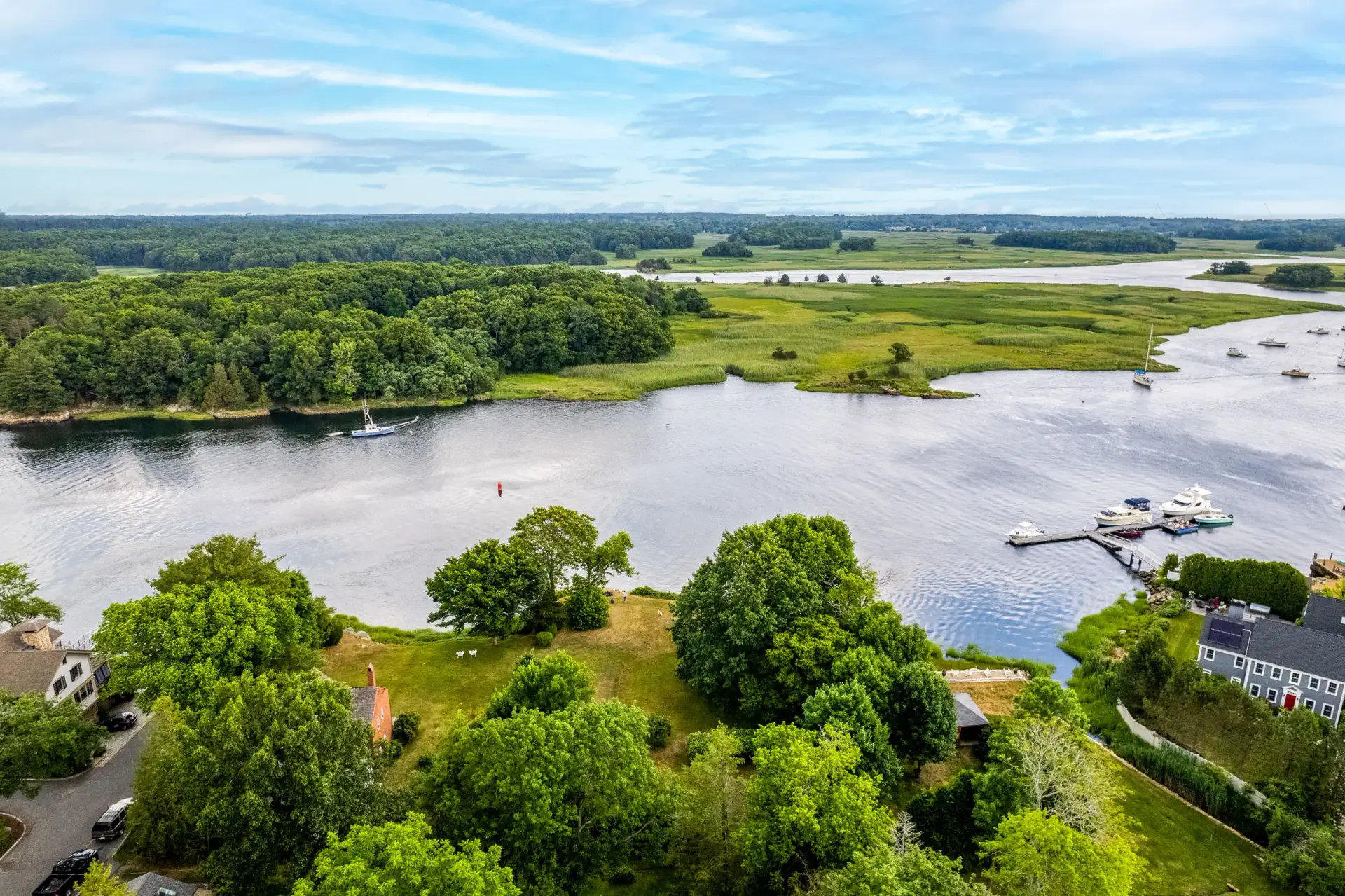 Aerial view of a serene waterway in Massachusetts with lush green landscapes, boats docked at a marina, and residential homes nearby, highlighting the natural beauty ideal for real estate marketing.