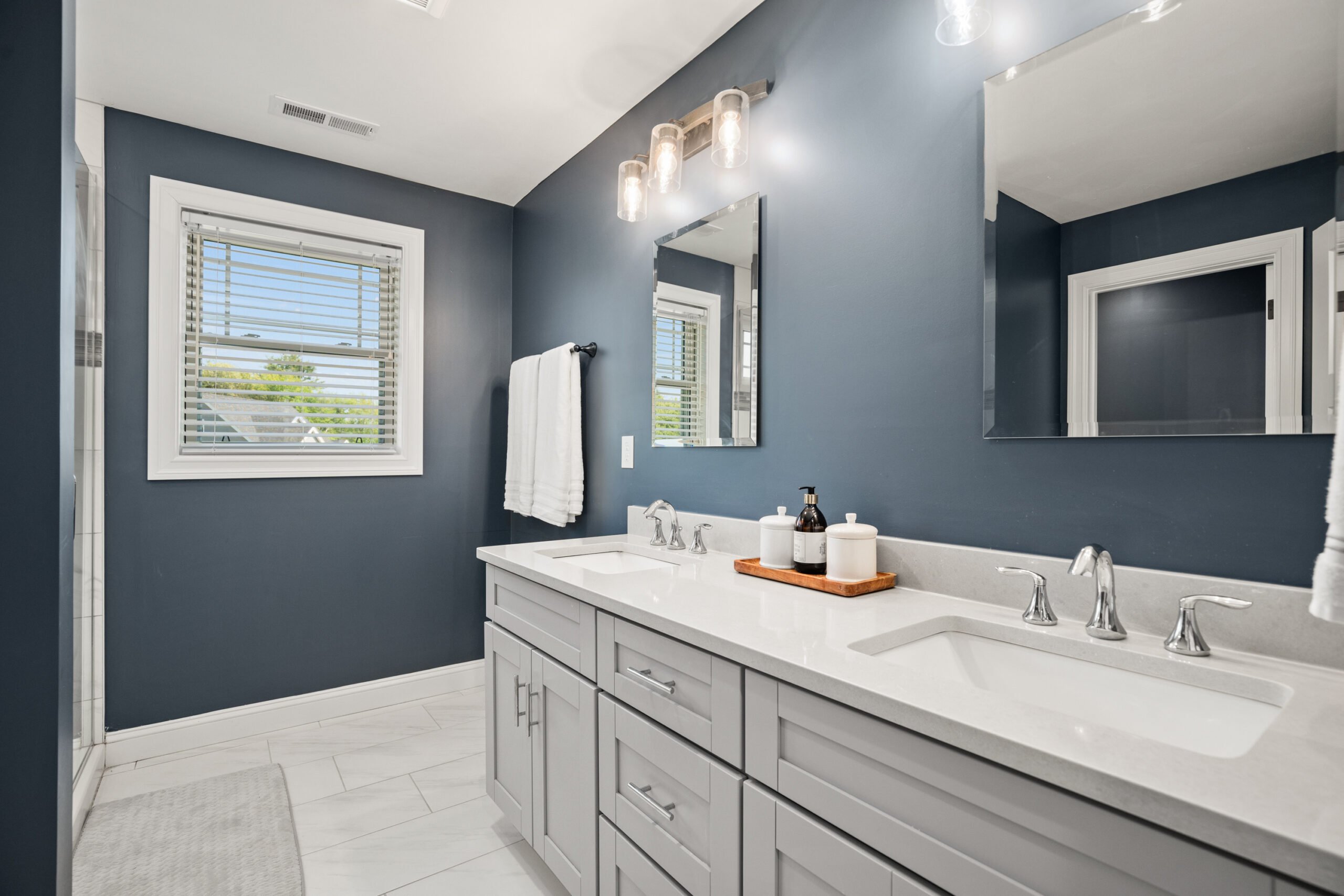 Modern bathroom with navy blue walls, double vanity sink, stylish fixtures, and natural light from a window, showcasing professional real estate photography for property listings.