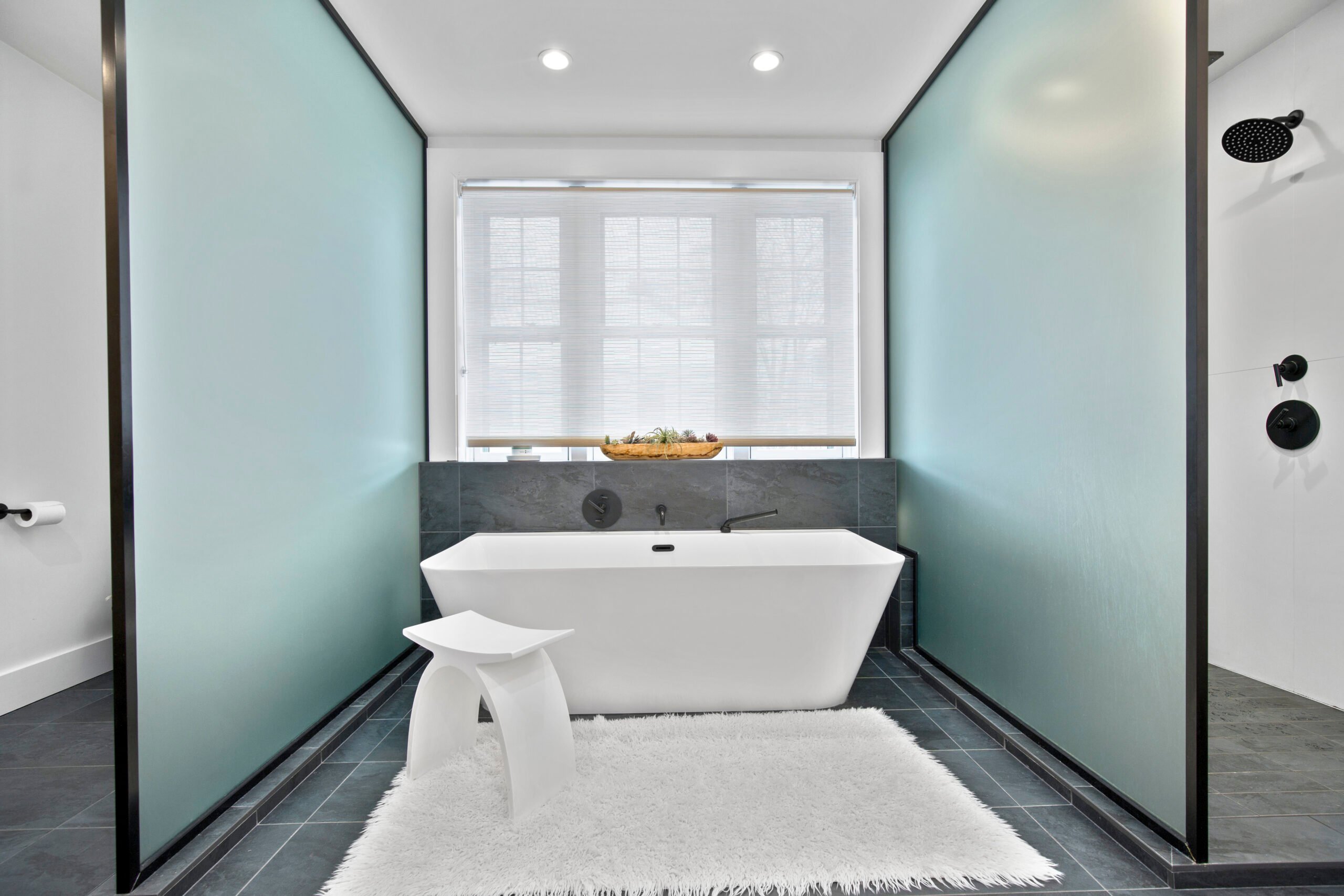 Modern bathroom featuring a freestanding white tub, sleek black fixtures, frosted glass partitions, and a minimalist design with a plush white rug.